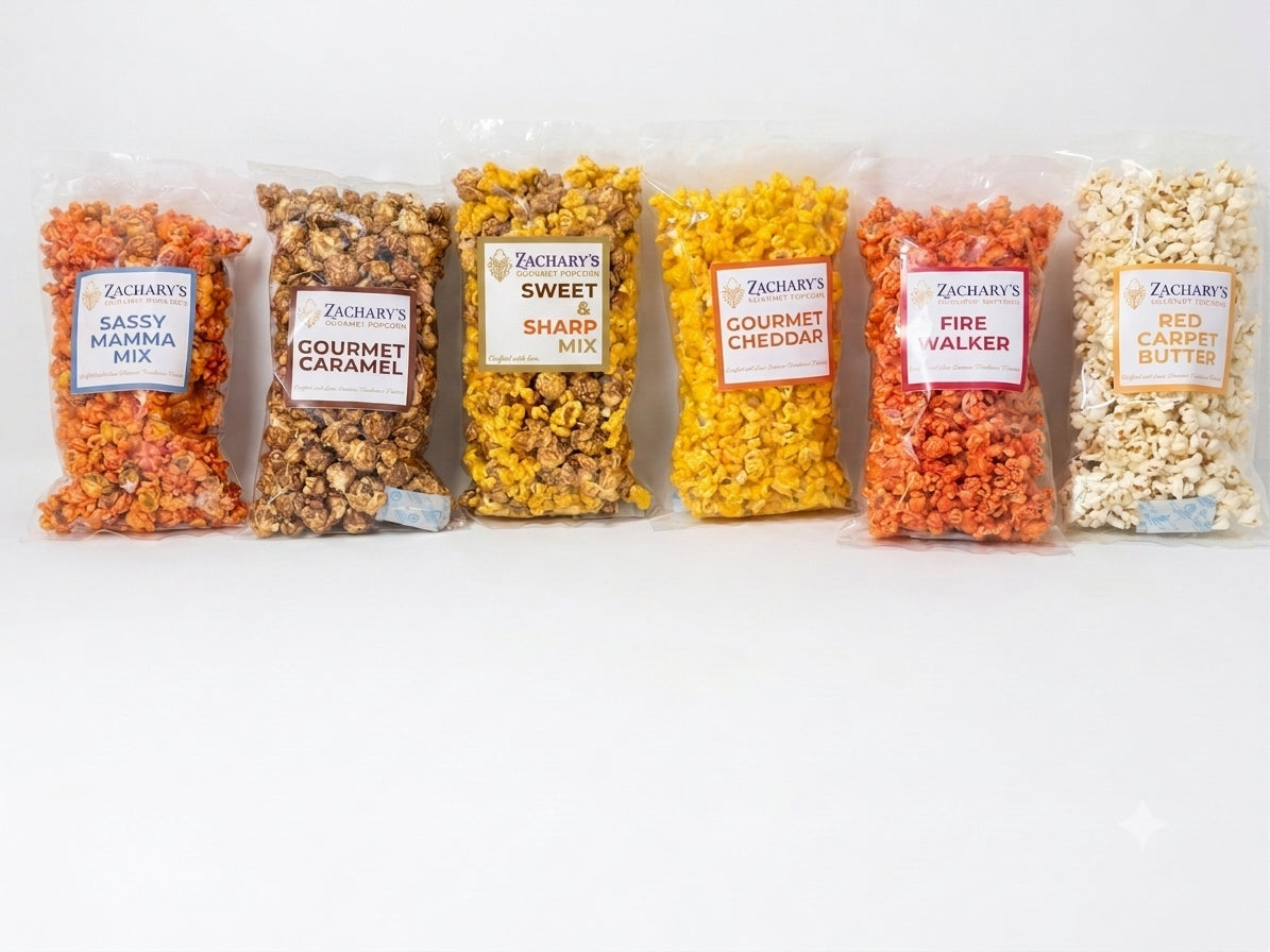 Six clear plastic bags of Zachary's gourmet popcorn mixes lined up on a white background. The flavors shown are Sassy Mama Mix, Gourmet Caramel, Sweet & Sharp Mix, Gourmet Cheddar, Fire Walker, and Red Carpet Butter.