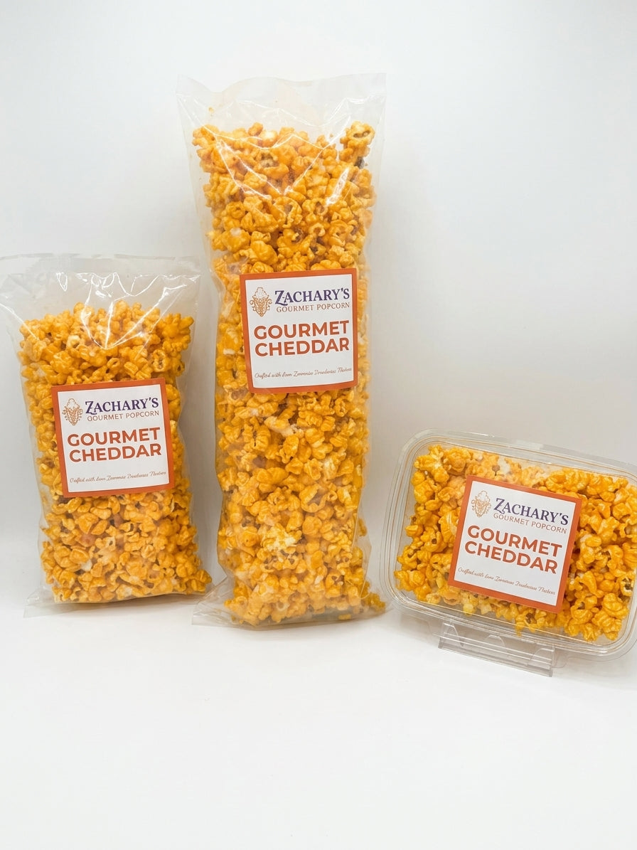 Three packages of Zachary's Gourmet Cheddar popcorn on a clean white background, including two clear bags and one clear plastic tub.